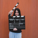 A young girl holding a movie clapboard in front of an orange background. Movie film director concept.