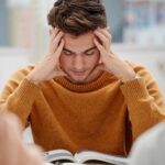 un estudiante leyendo un libro escolar, concentrado y sujetándose la cabeza con ambas manos.